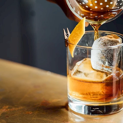 A whiskey cocktail being poured into a rocks glass with ice, garnished with an orange twist and a small clip, stylish bar scene.