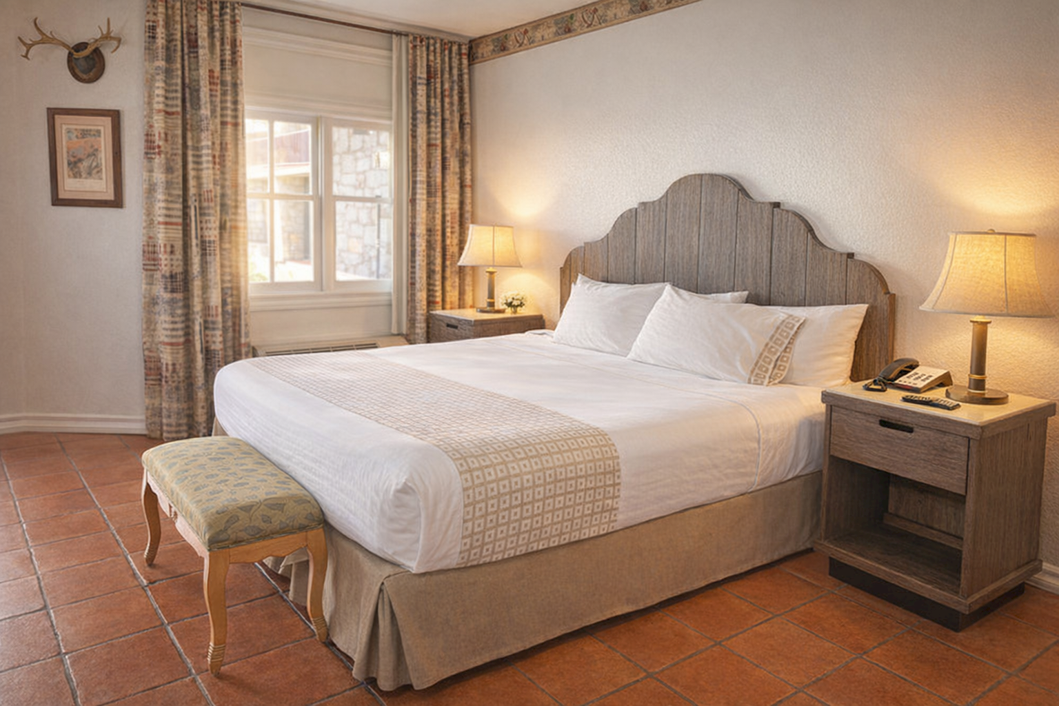 A cozy hotel room with a large bed, upholstered headboard, two nightstands with lamps, a bench, window with curtains, and terracotta tile floor.