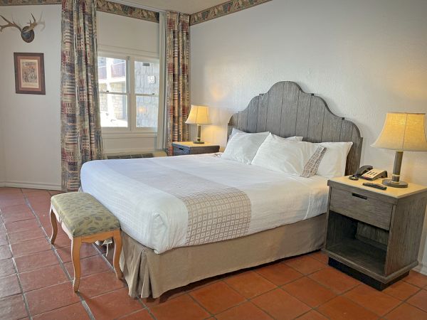 The image shows a cozy bedroom with a large bed, pillows, two bedside tables with lamps, a window, and decorative curtains.