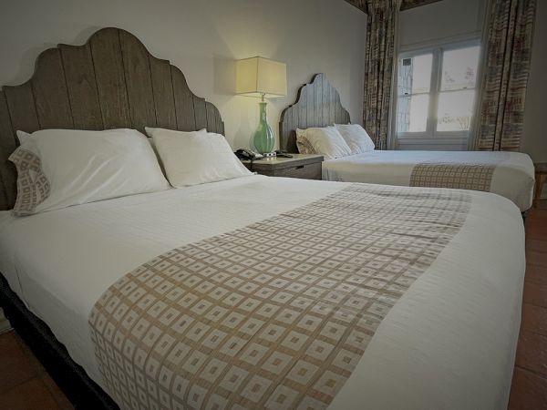 The image shows a hotel room with two double beds, each with white sheets and patterned blankets, a bedside table, lamp, and a window.