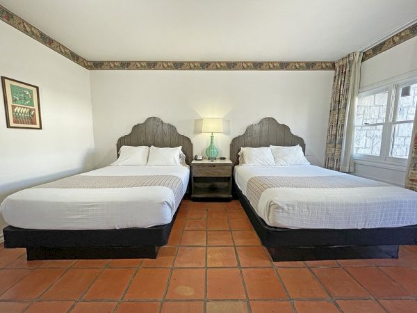 A hotel room with two beds, white bedding, a lamp on a nightstand, artwork on the wall, and a window with curtains.