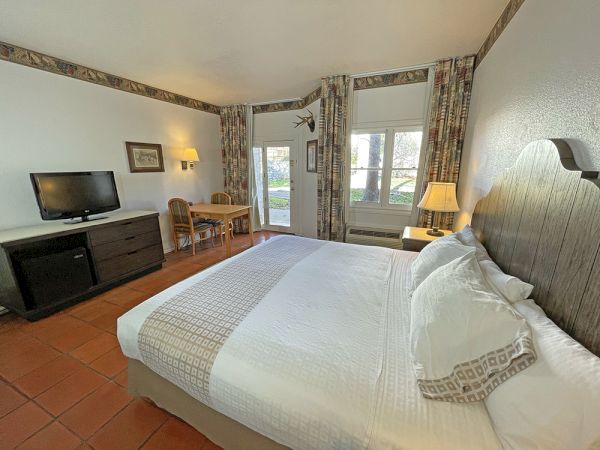 The image shows a hotel room with a bed, TV, desk, lamp, and large windows with curtains, creating a cozy atmosphere.