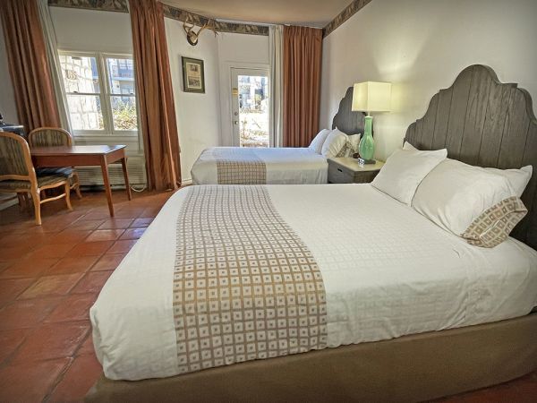 The image shows a hotel room with two neatly made beds, a desk and chair, curtains, and a lamp on the bedside table.