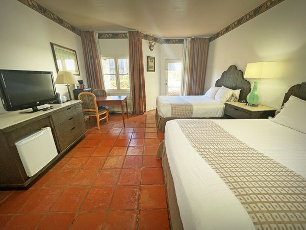 A hotel room with two beds, a TV, a table with chairs, a lamp, and a mini-fridge on a tiled floor, near a window with curtains.
