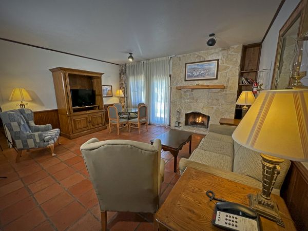 A cozy living room with a fireplace, armchairs, a wooden cabinet with a TV, and a dining table, illuminated by warm lighting.