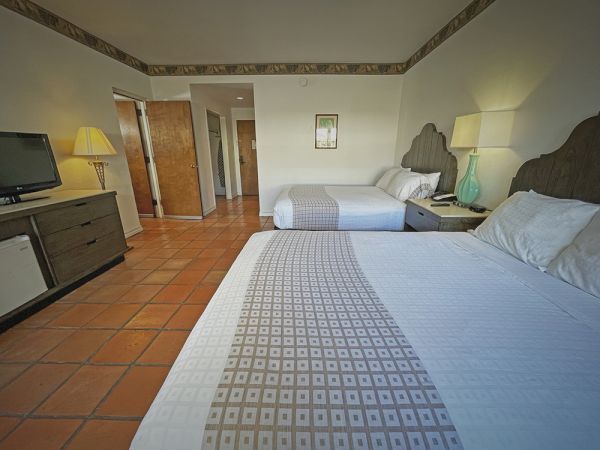 The image shows a hotel room with two beds, lamps, a TV, and a dresser on tiled flooring, featuring a cozy and simple decor.