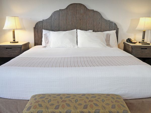 A neatly made bed with white linens, two pillows, a wooden headboard, and two bedside tables with lamps on each side, and a bench at the foot.