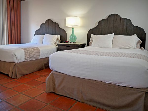 The image shows a cozy hotel room with two double beds, a wooden headboard, a bedside lamp on a table, and terracotta tile flooring.