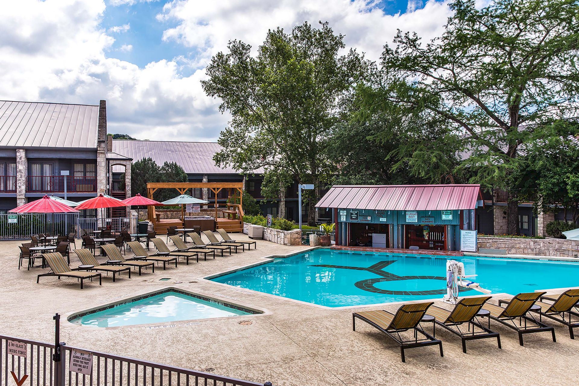 An outdoor hotel pool with turquoise water, sun loungers, red umbrellas, and low buildings under a cloudy blue sky.