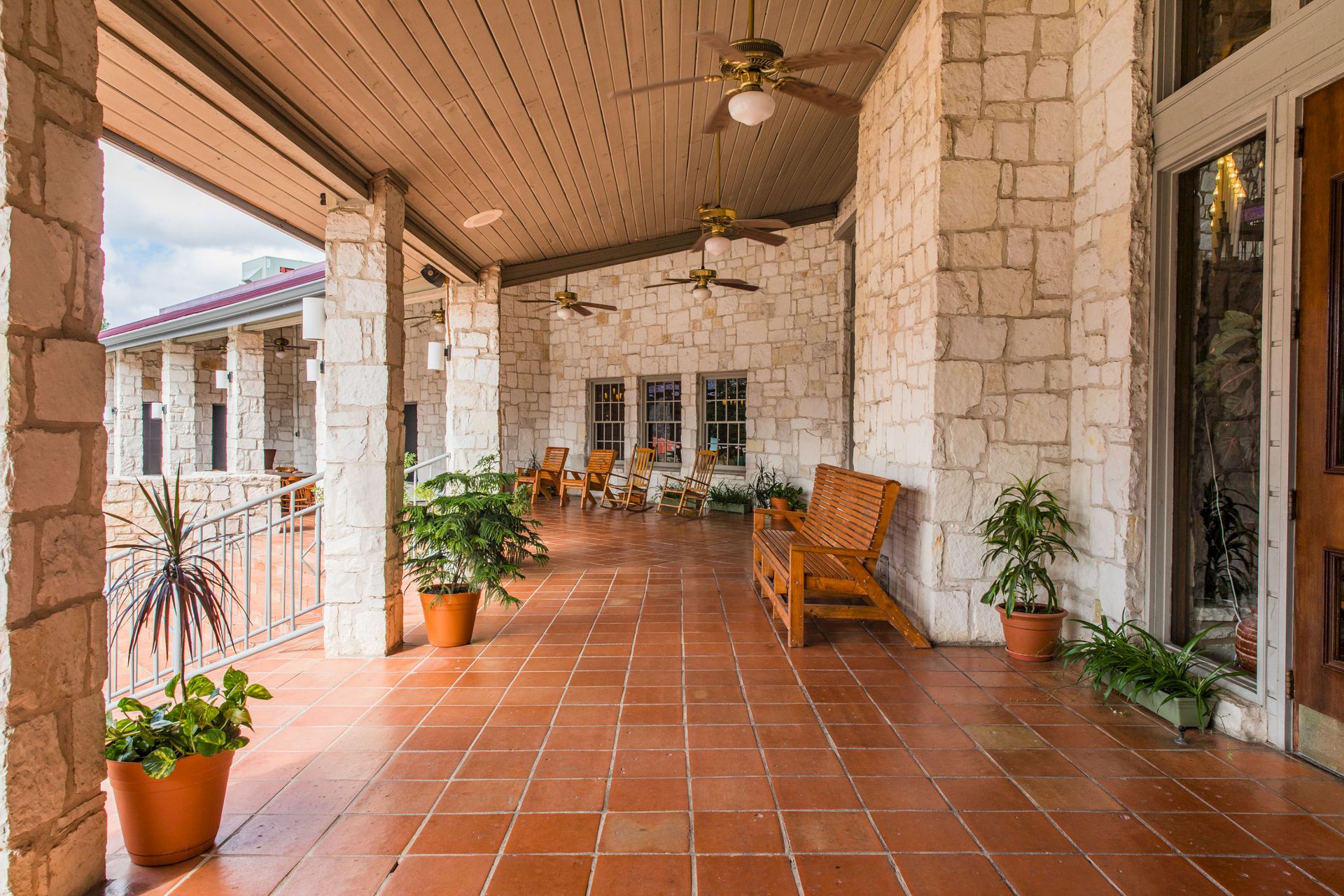 A covered stone patio with tiled floor, ceiling fans, wooden chairs, and potted plants lining the walkway.