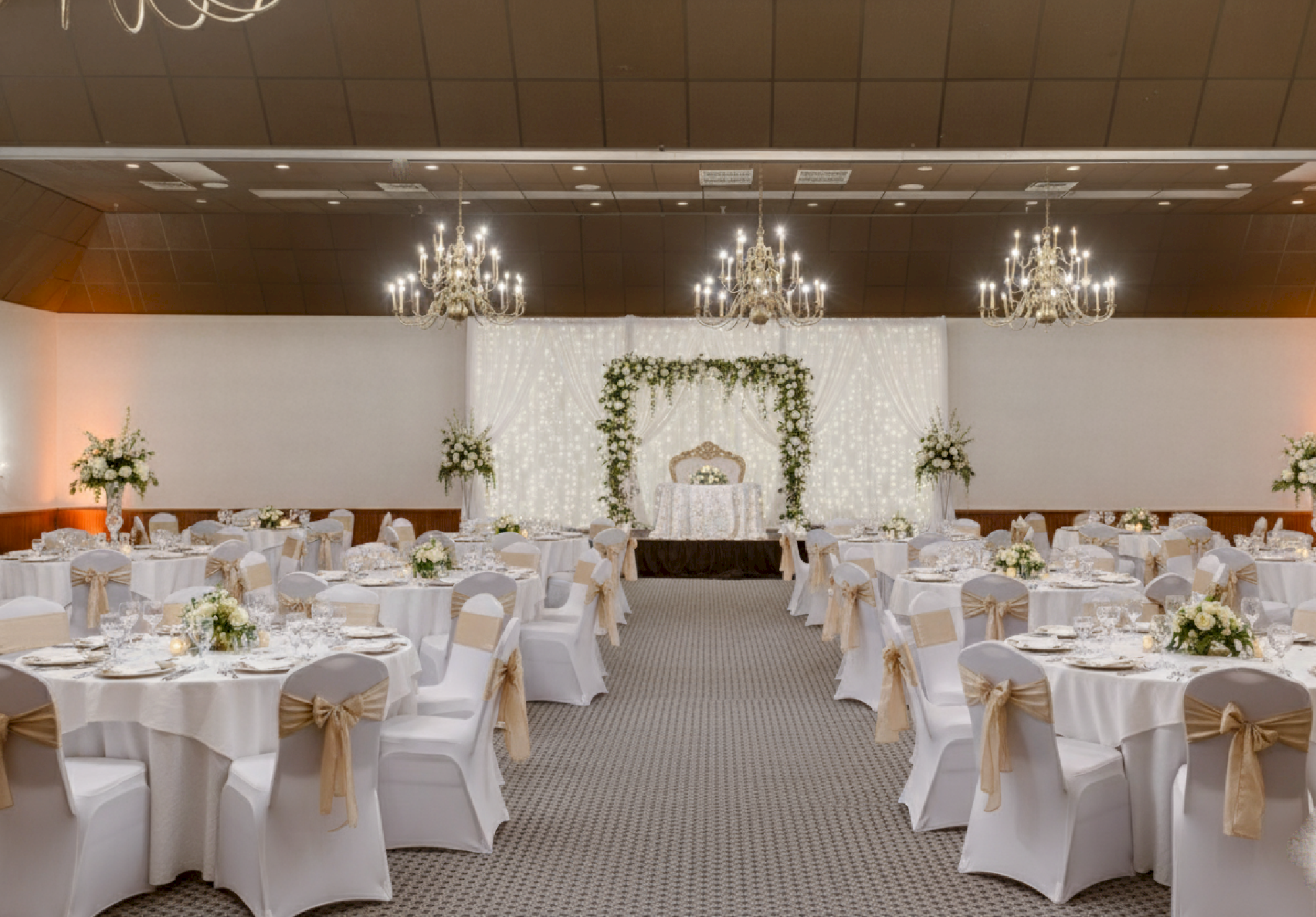 The image shows a decorated banquet hall with round tables, white chairs, floral centerpieces, chandeliers, and an elegant backdrop.