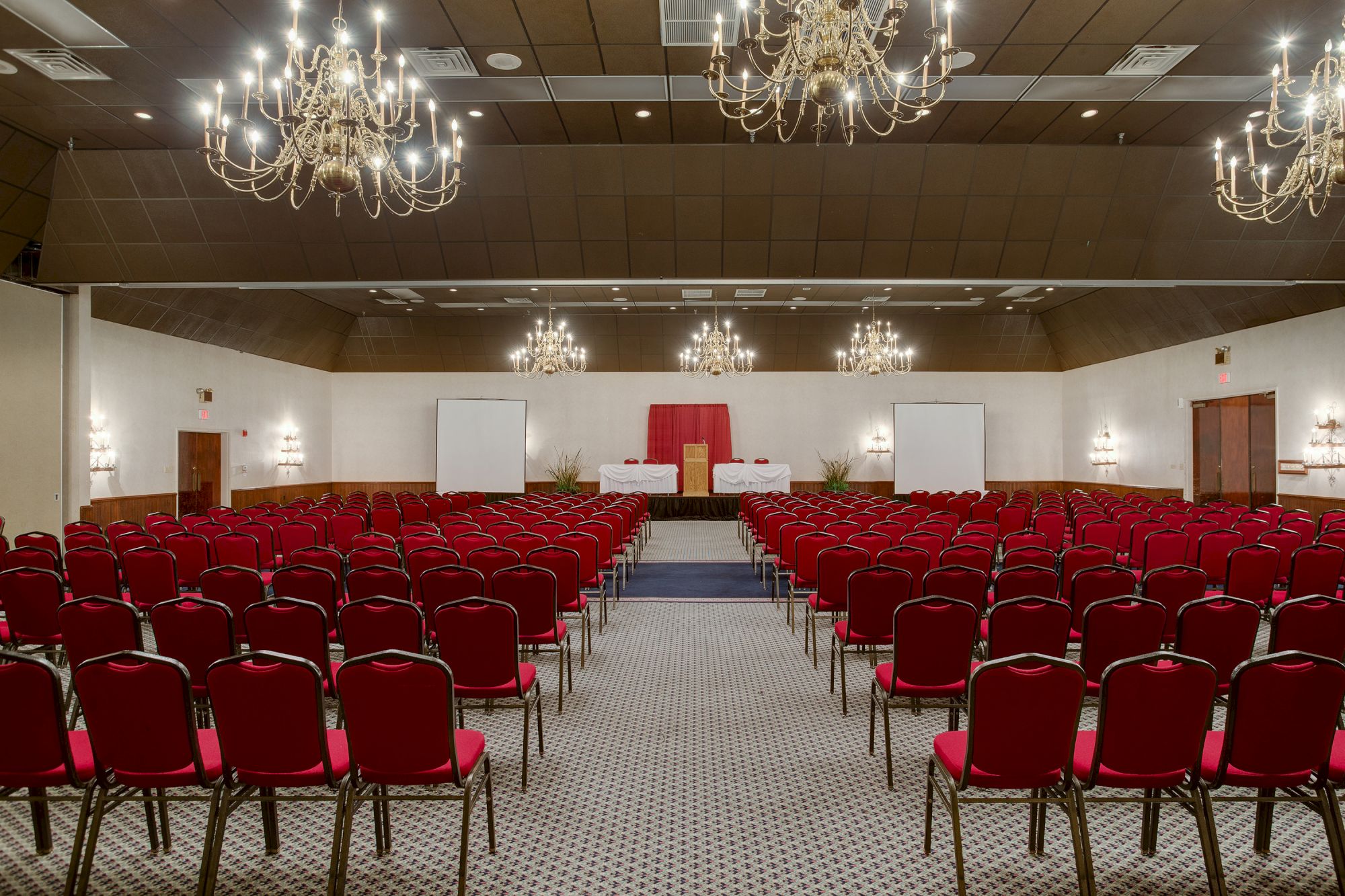 The image shows a large conference room with red chairs arranged in rows, chandeliers overhead, and a stage with a podium at the front.