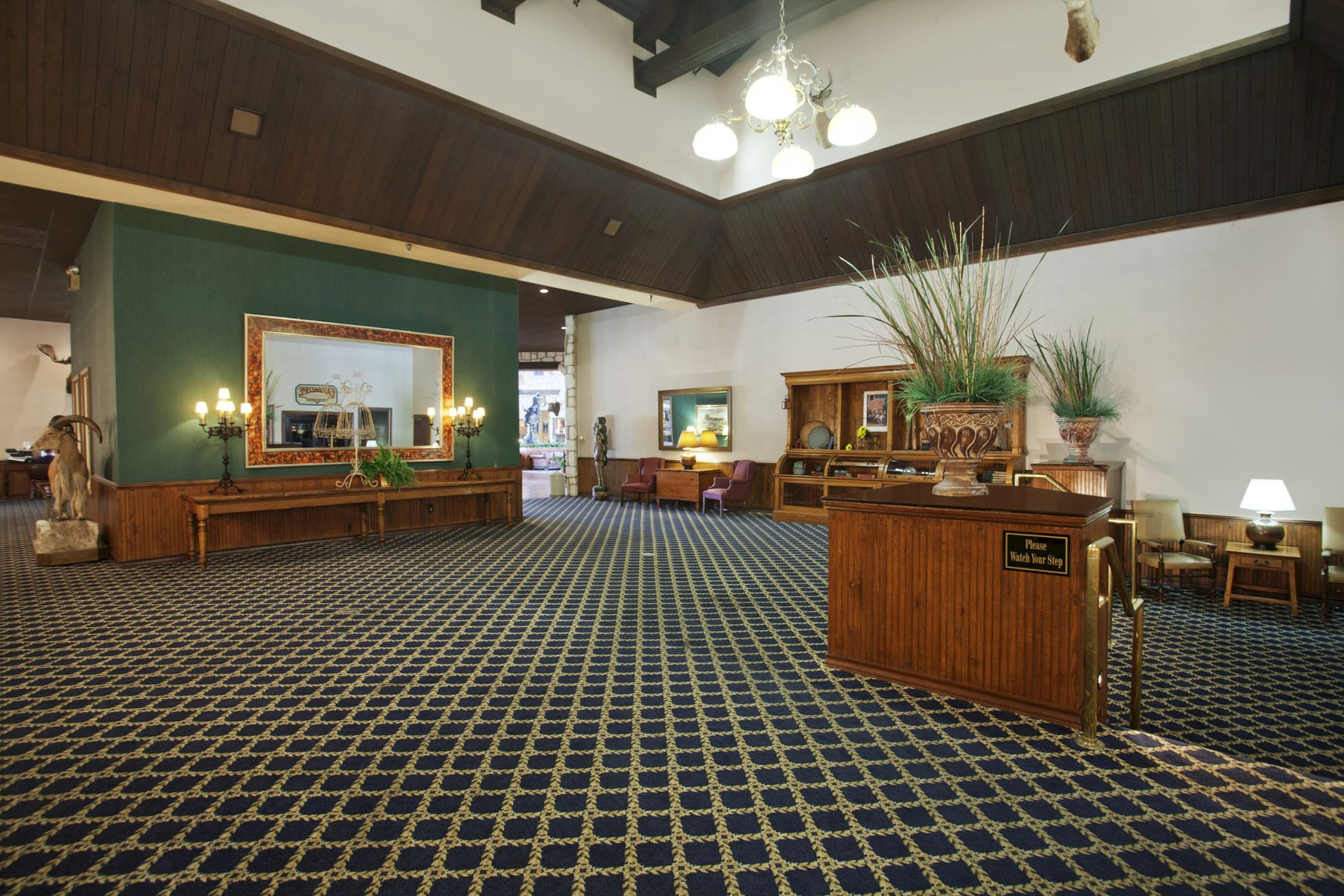 The image shows a spacious, carpeted lobby with wooden furniture, art pieces on the walls, large mirrors, and decorative lights.