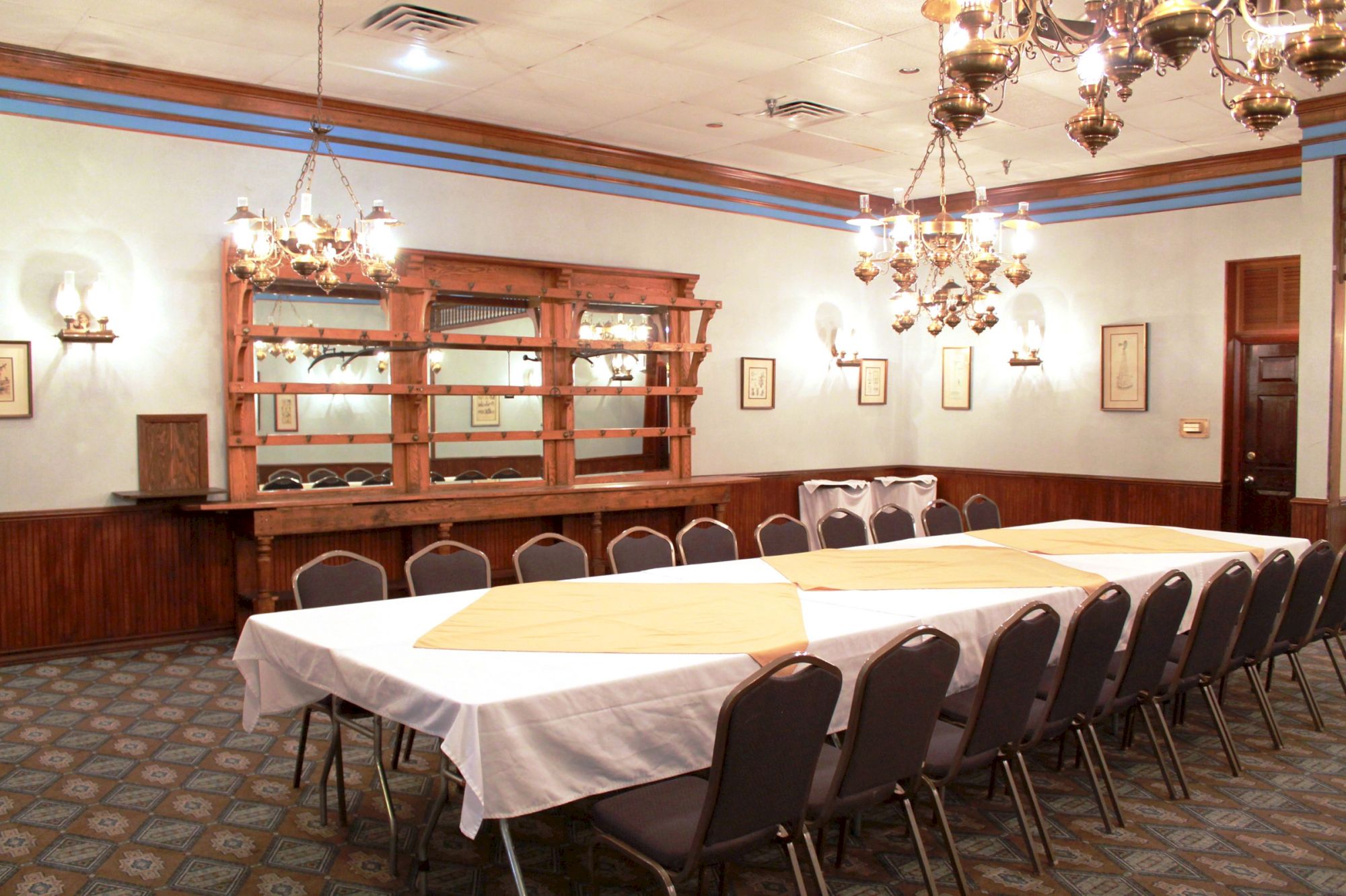 A dining room with a long table covered with a white cloth, surrounded by chairs, chandeliers above, and a wooden cabinet in the background.