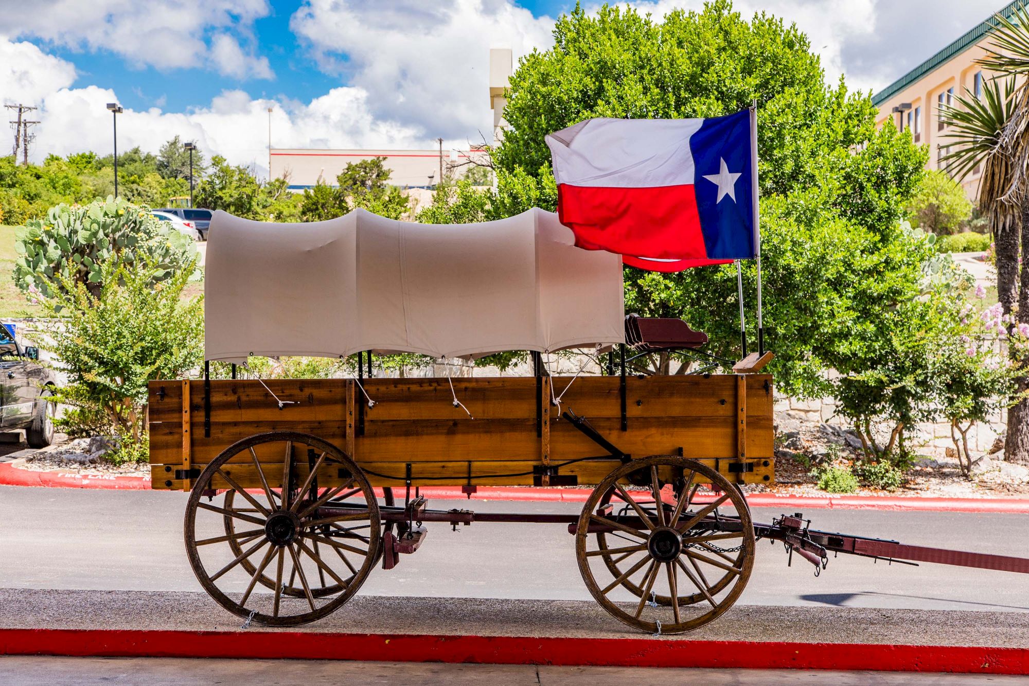 A covered wooden wagon with a Texas flag is displayed outdoors on a sunny day, surrounded by greenery and buildings.