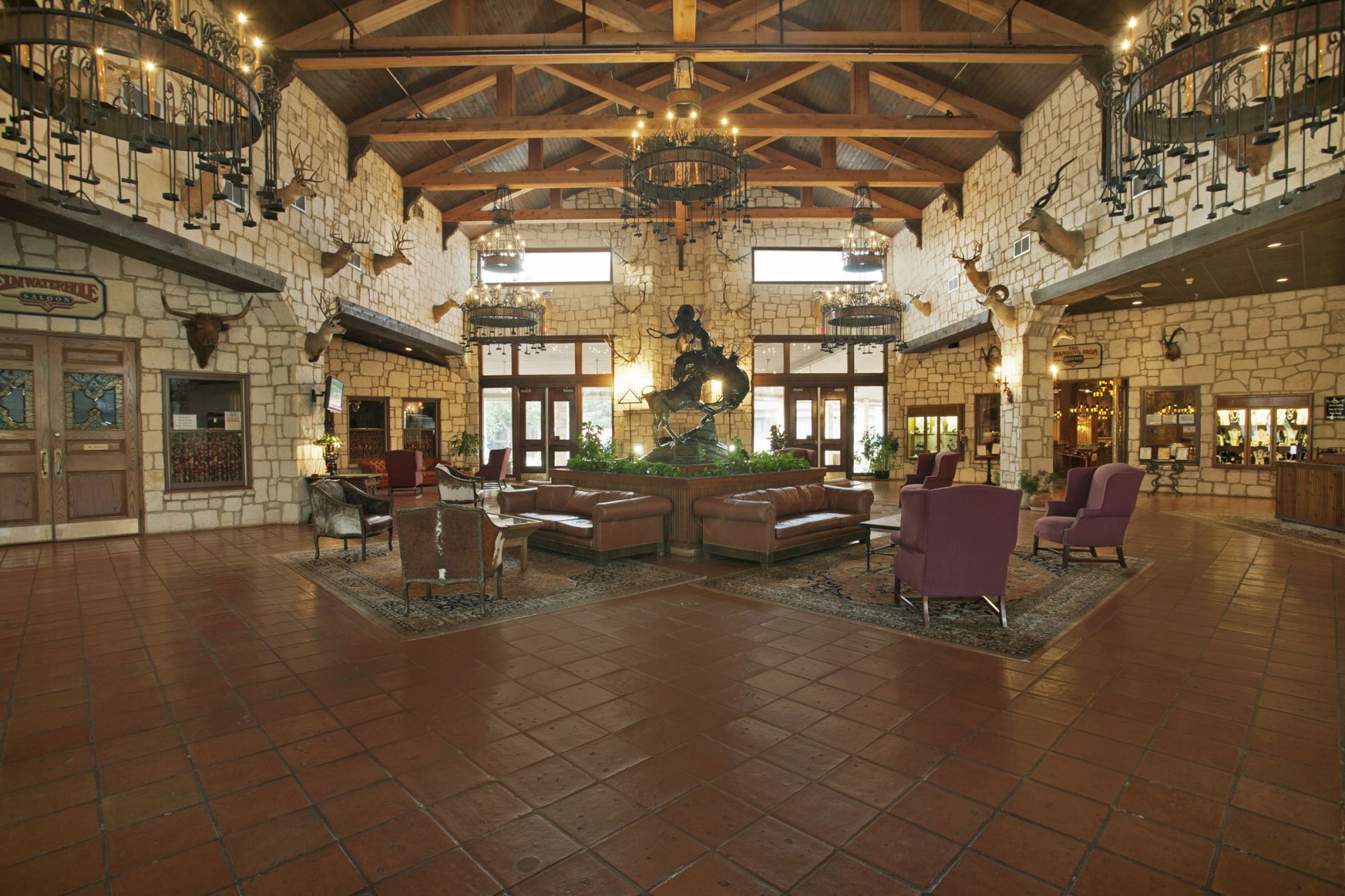 A spacious, rustic lobby with stone walls, chandeliers, cozy seating, and a central plant display under a cathedral ceiling.