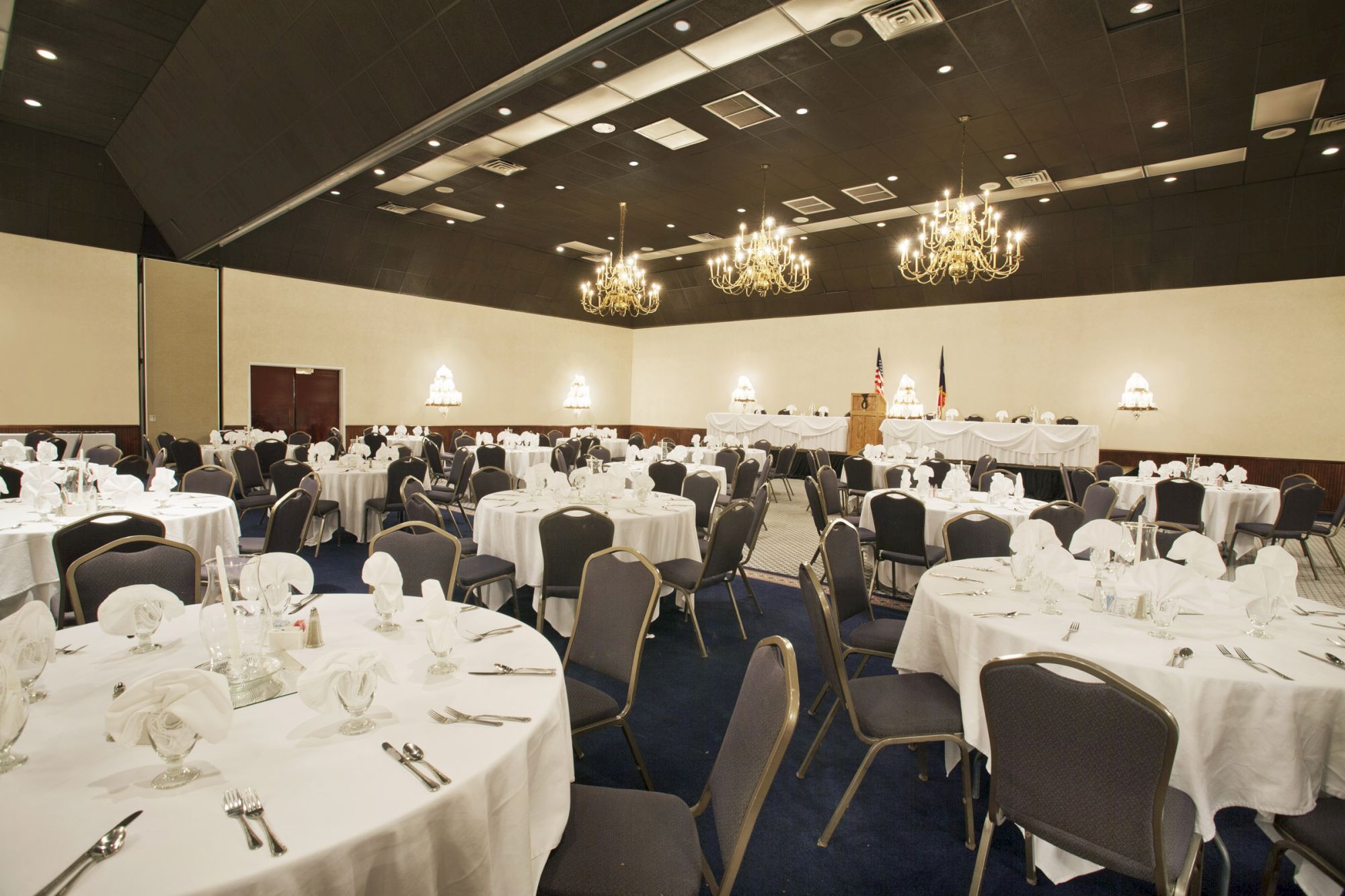 The image shows a banquet hall with round tables set for a formal event, with white tablecloths, chairs, and chandeliers overhead.