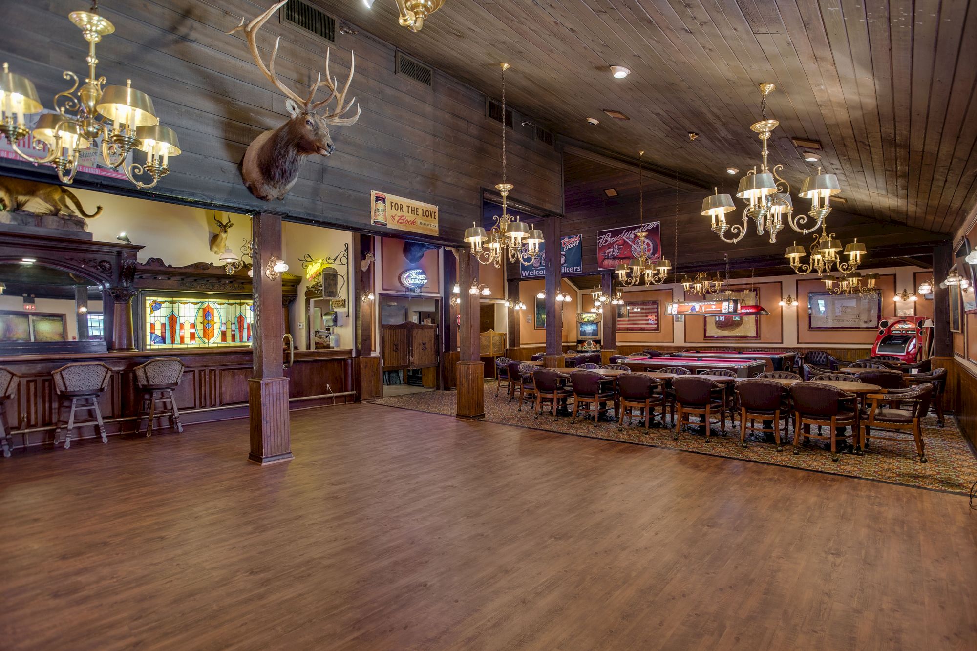 The image shows a spacious bar with chandeliers, a mounted deer, wooden decor, a long bar, and seating area in a rustic style.