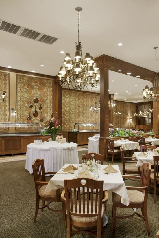 The image shows an elegant restaurant interior with wooden decor, chandeliers, round tables with white tablecloths, and upholstered chairs.