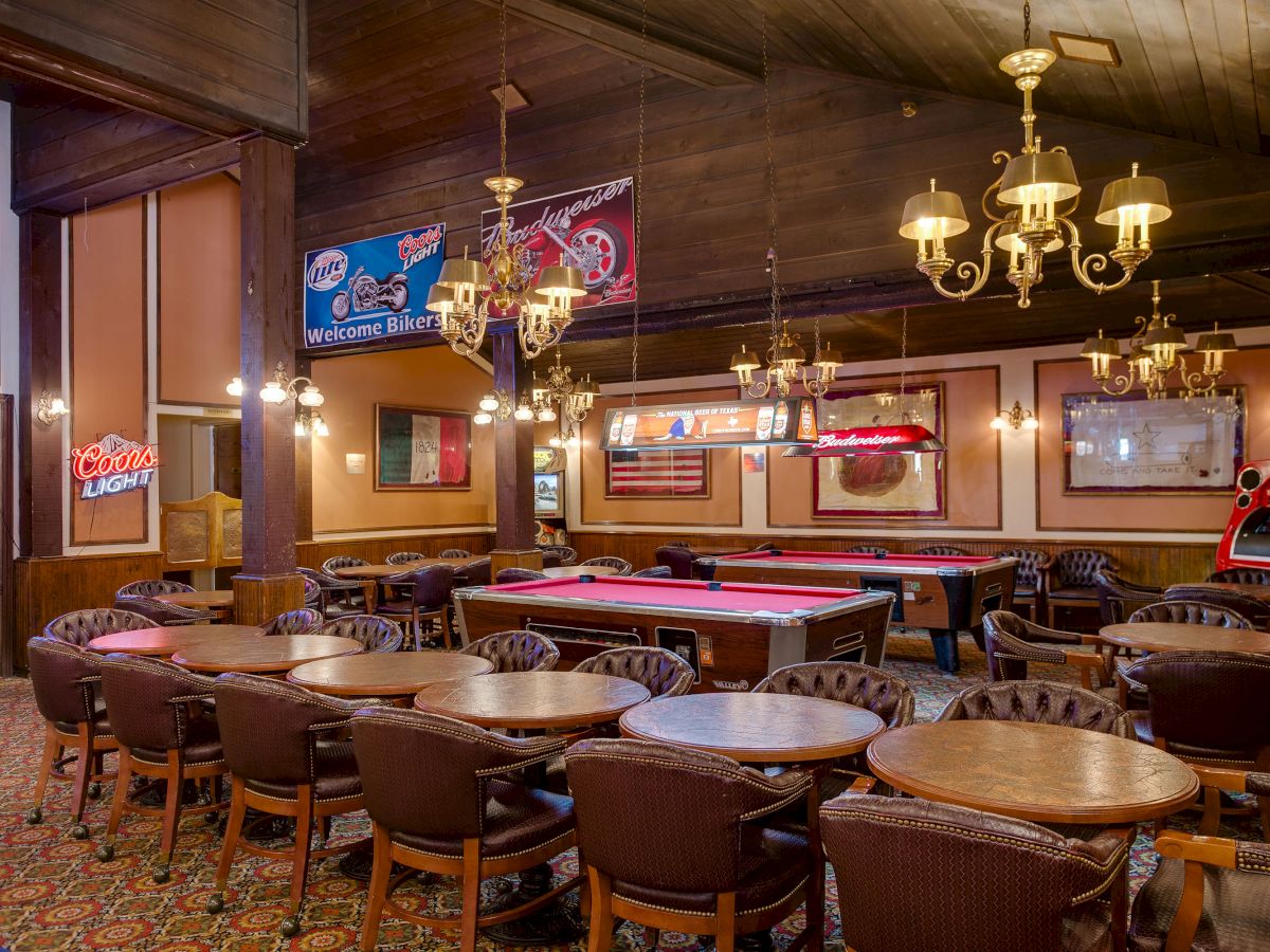 The image shows an empty bar area with pool tables, round wooden tables, chairs, chandeliers, and neon signs on the walls.