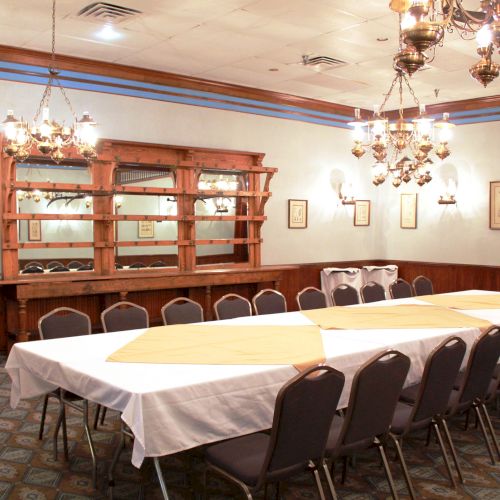 A dining room with a long table, chairs, chandeliers, and a wooden bar with shelves. The table is set with white and yellow tablecloths.