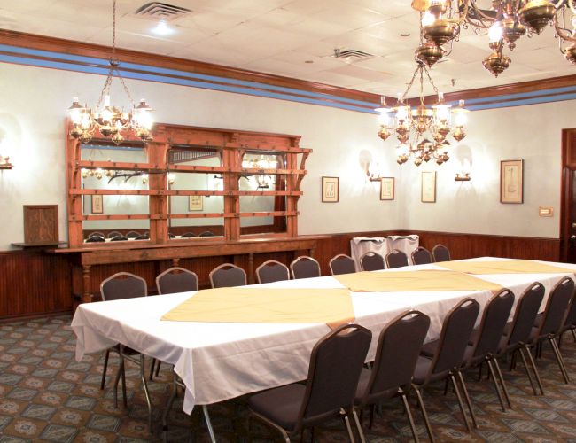 A dining room with a long table, chairs, chandeliers, and a wooden bar with shelves. The table is set with white and yellow tablecloths.