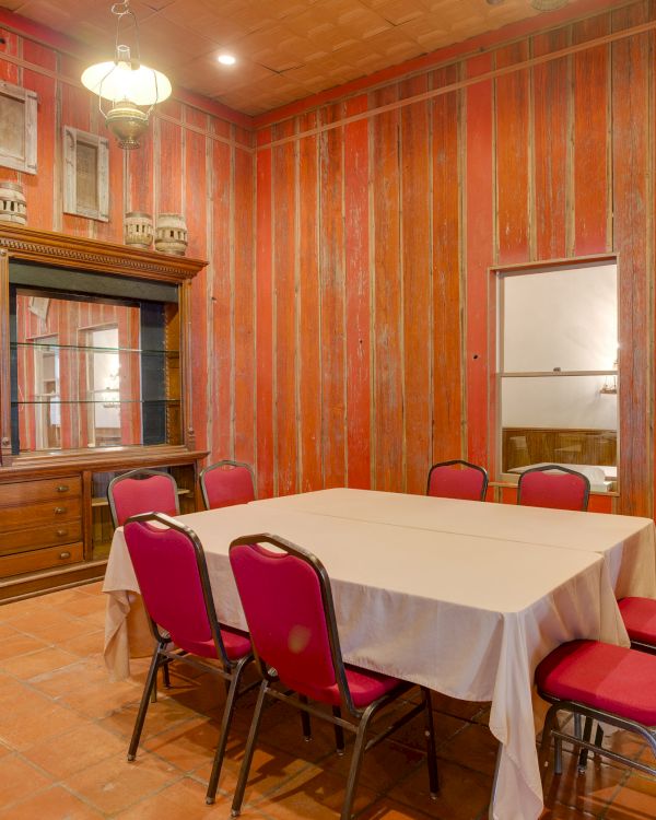 The image shows a rustic room with wooden walls, a table covered with a cloth, red chairs, and display shelves.