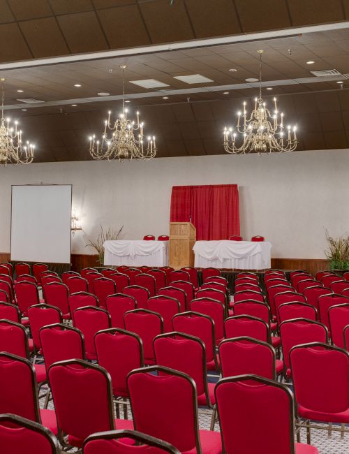 An empty conference hall with rows of red chairs, chandeliers, two screens, and a podium with red drapes at the front.