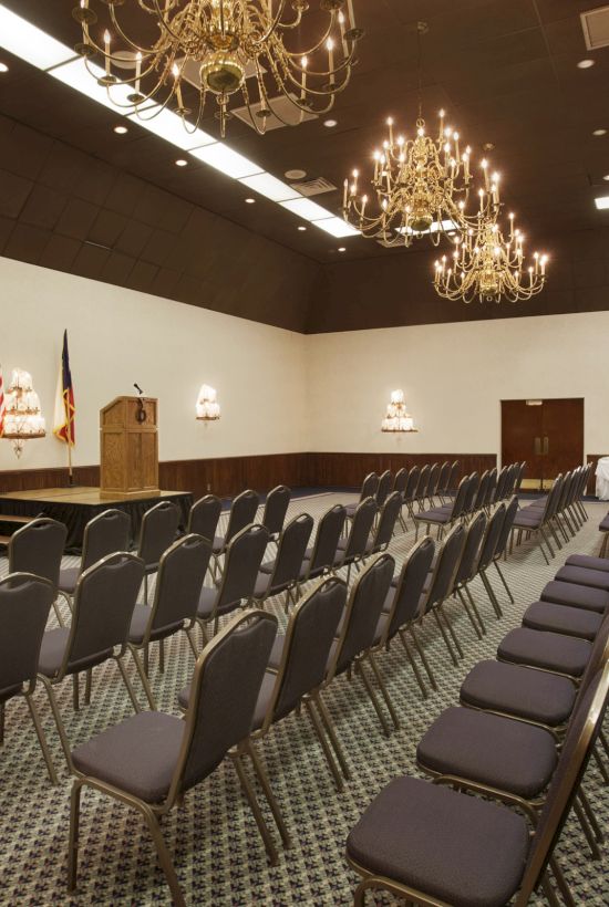The image shows a conference room with rows of empty chairs, chandeliers, and a podium, likely set up for a meeting or event.