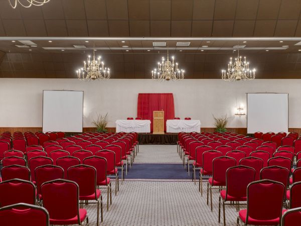An empty conference room with red chairs, two projection screens, chandeliers, and a podium at the front on a stage with a red backdrop.