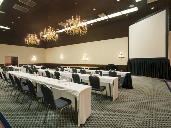 A conference room with rows of tables and chairs, chandeliers, wall lights, and a large projector screen at the front of the room.