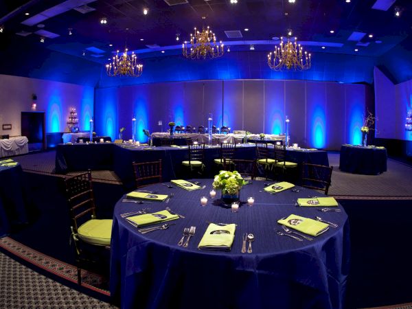 The image shows an elegantly set event space with blue lighting, round tables with green napkins, and floral centerpieces.