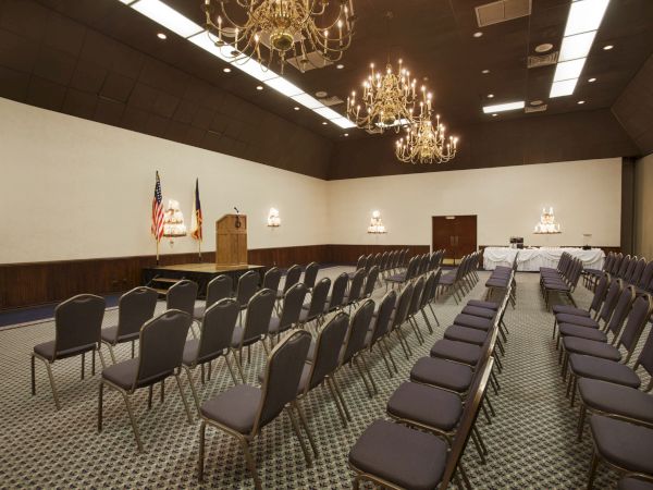 An empty conference room with rows of chairs, a podium with flags, chandeliers, and tables at the back, set for an event or meeting.