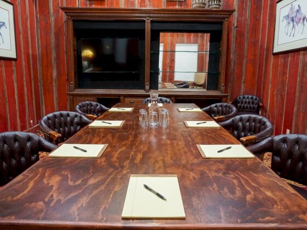 A conference room with a large wooden board table, leather chairs, notepads, pens, and a cabinet mirror on red striped walls.