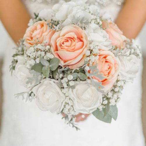 A bride holding a bouquet of peach and white roses with greenery, wearing a lace wedding dress.