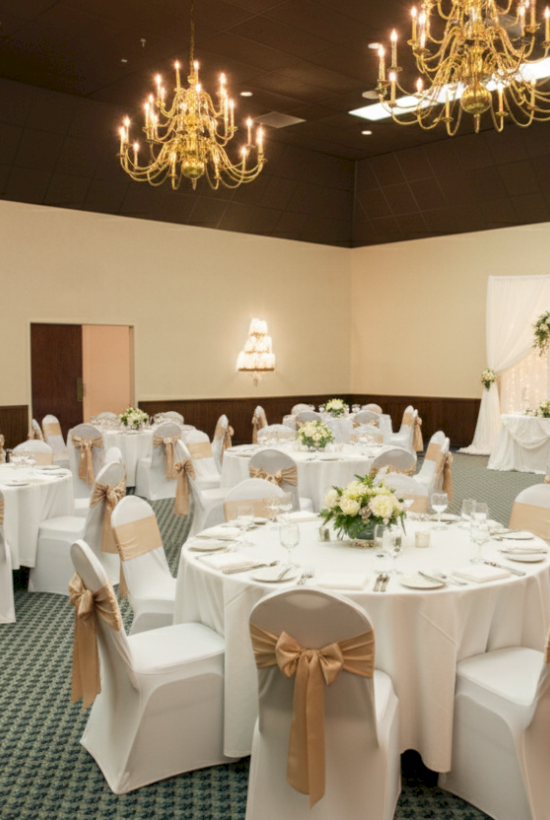 The image shows a decorated banquet hall with round tables, white chairs with bows, floral centerpieces, and chandeliers overhead.