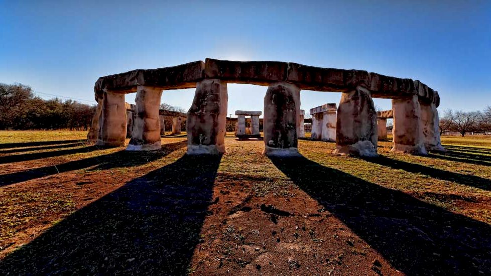 A row of weathered stone slabs and arches at a historical memorial site under a clear blue sky.