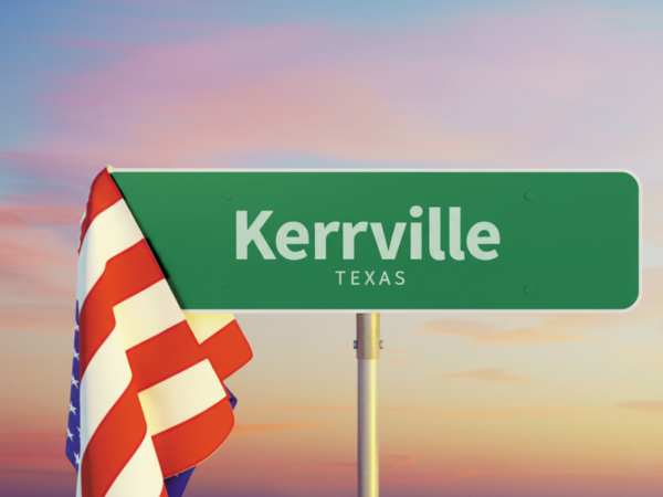 A green road sign reads "Kerrville, Texas" with an American flag draped beside it, set against a sky at sunset.