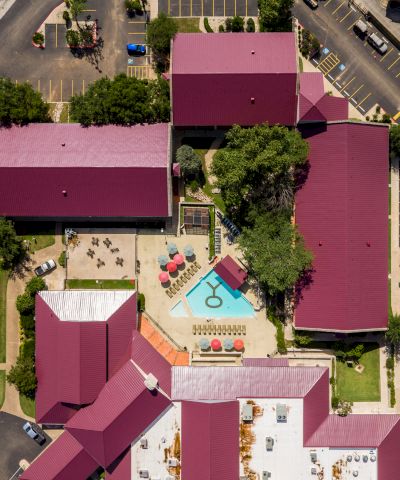An aerial view of a colorful resort-like complex with pink rooftops, a central pool area, and manicured courtyards surrounded by streets.
