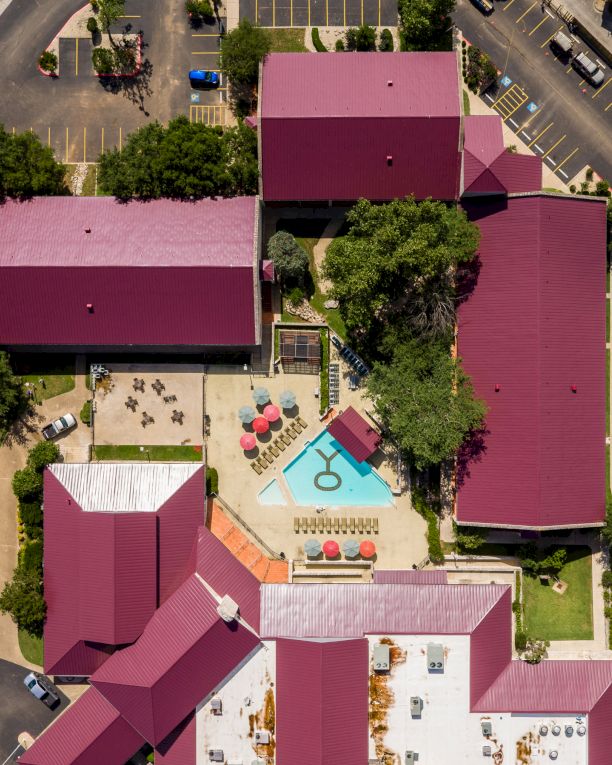 An aerial view of a colorful resort-like complex with pink rooftops, a central pool area, and manicured courtyards surrounded by streets.
