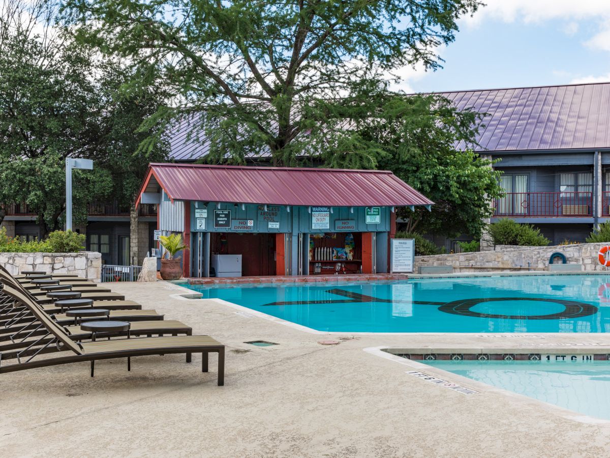 The image shows a pool area with lounge chairs, a poolside bar with a red roof, and buildings in the background under a partly cloudy sky.