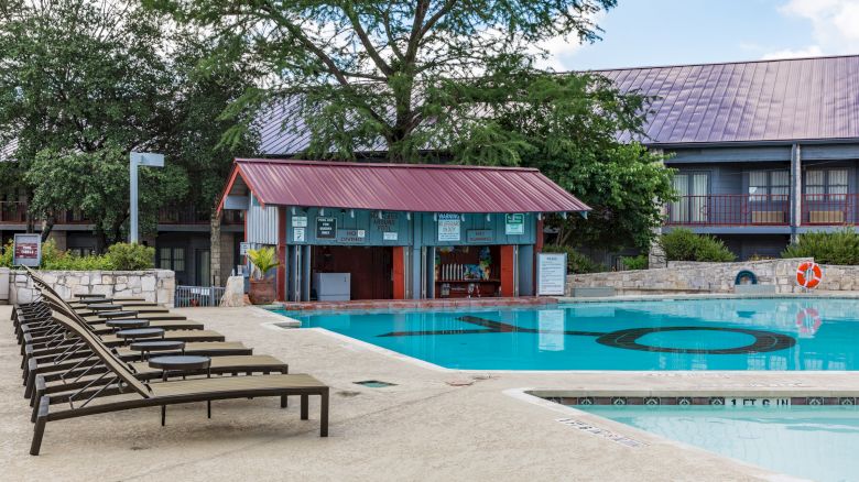 The image shows a pool area with lounge chairs, a poolside bar with a red roof, and buildings in the background under a partly cloudy sky.