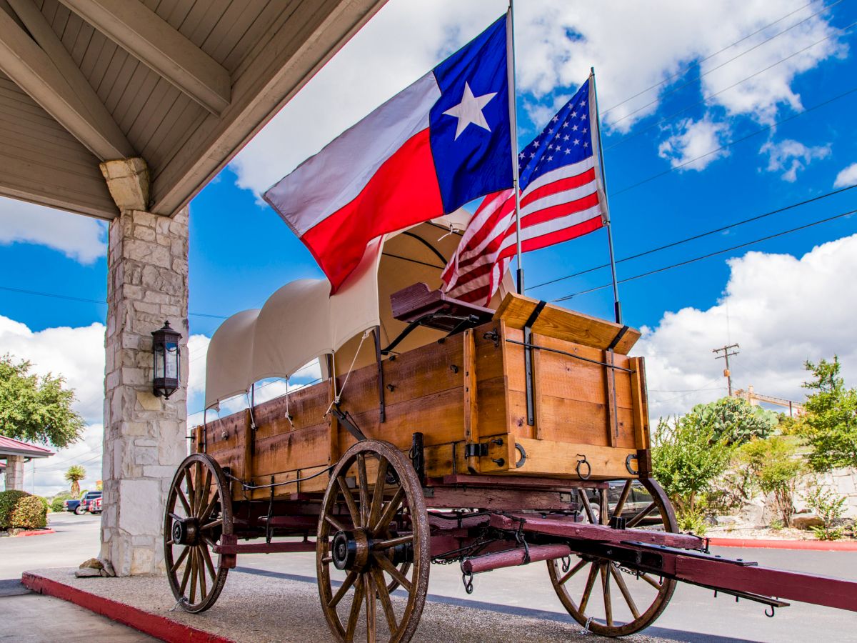 A covered wagon is parked under a roof with Texas and U.S. flags flying above, set against a blue sky with clouds.
