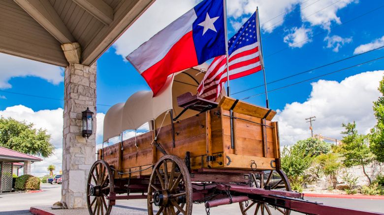 A covered wagon is parked under a roof with Texas and U.S. flags flying above, set against a blue sky with clouds.