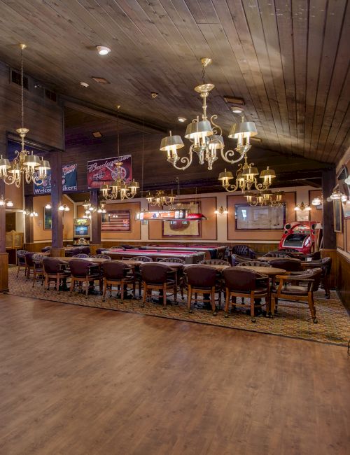 A large, rustic bar with chandeliers, wooden floors, tables, deer mounts, and neon signs, giving a cozy and traditional atmosphere.