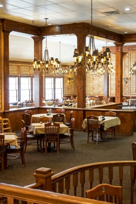 A warm, wood-paneled restaurant dining room with long tables and ornate chandeliers hanging overhead, lit for diners.