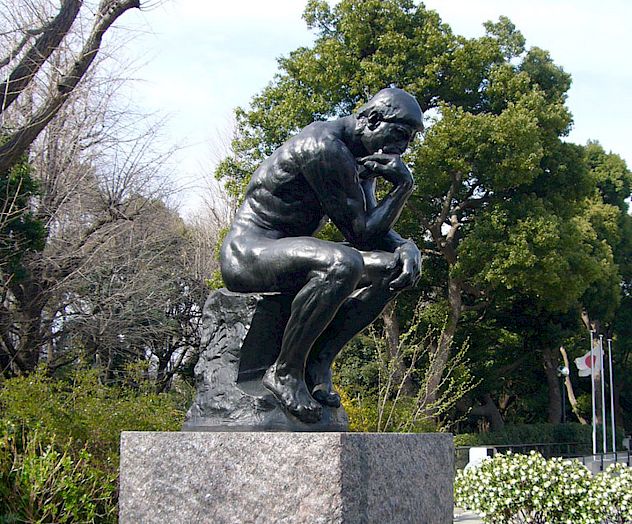 This image shows a statue of a person sitting and deep in thought, placed on a stone pedestal outdoors amidst trees and greenery.