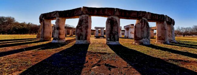 The image shows a replica of Stonehenge, with large stone structures casting long shadows on the ground under a clear blue sky.