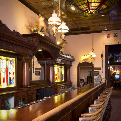 Vintage bar with ornate wooden counter, stained glass accents, and classic pendant lighting, creating an inviting atmosphere.
