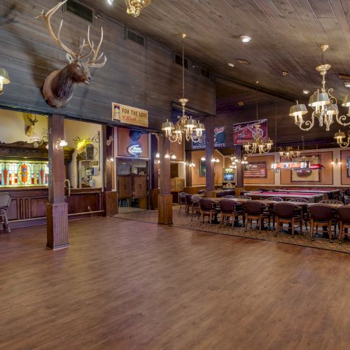 A rustic bar interior with wooden floors, chandeliers, bar seating, and mounted animal heads, creating a cozy, vintage atmosphere.
